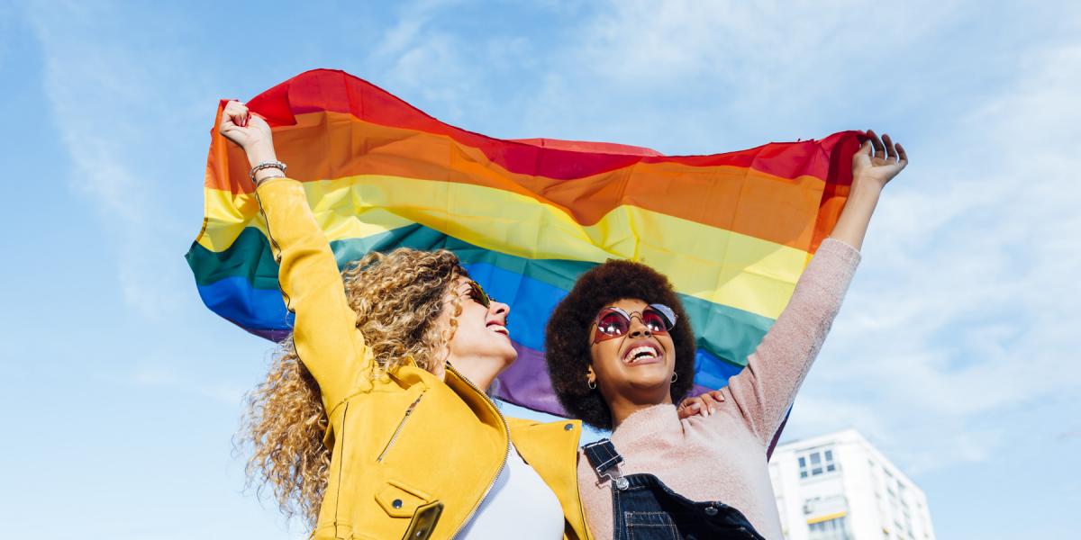 Two women holding the Pride flag over their heads, smiling 